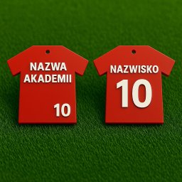 HexoCAD - Dwa czerwone breloczki w kształcie koszulek z białymi napisami 'Nazwa Akademii' i 'Nazwisko' z numerem 10, leżące na zielonej, teksturowanej powierzchni.