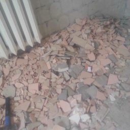ADRIAN ZIELIŃSKI - Stos potłuczonych, różowych i szarych płytek na podłodze w rogu pokoju, obok grzejnika i młotka. Widoczne ślady po demontażu płytek na ścianie.