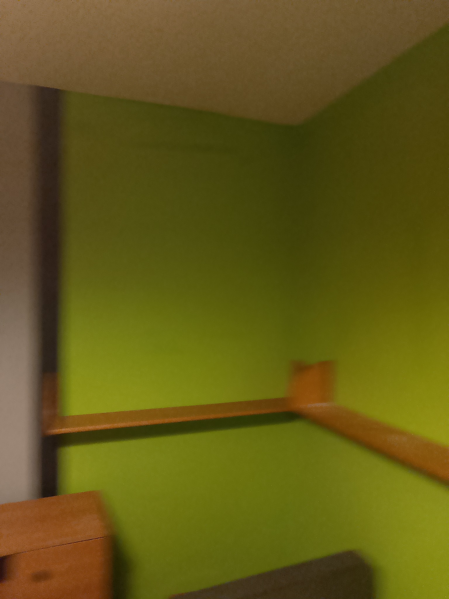 Fragment wnętrza z zieloną ścianą i drewnianą półką. Nierównomierne oświetlenie, rozmycie. Widoczny fragment mebla w dolnej części kadru.