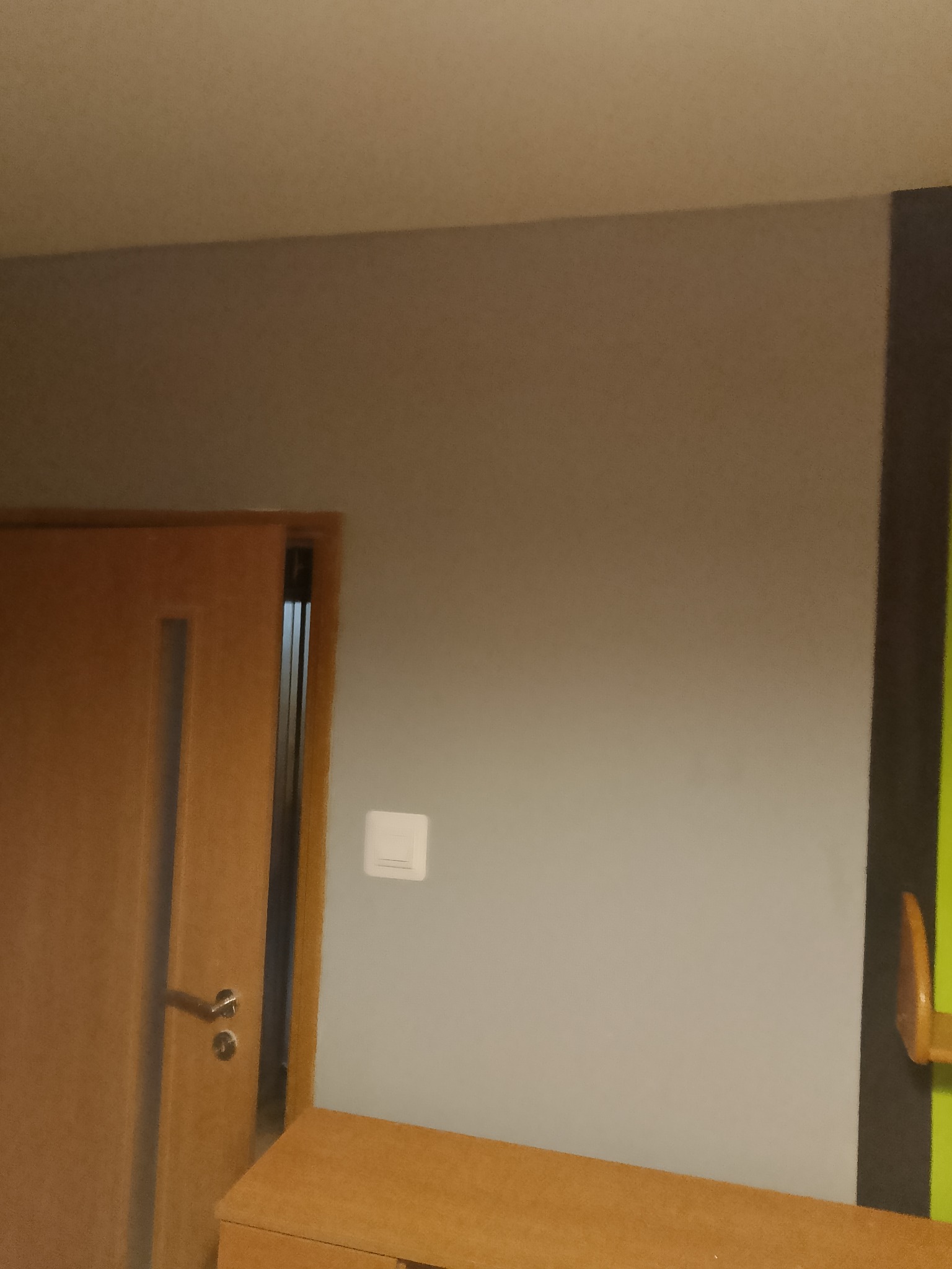 Szara ściana z drewnianymi drzwiami i szafką w pokoju. Widoczny włącznik światła i fragment zielonej ściany z czarną obwódką. Ujęcie z dołu.