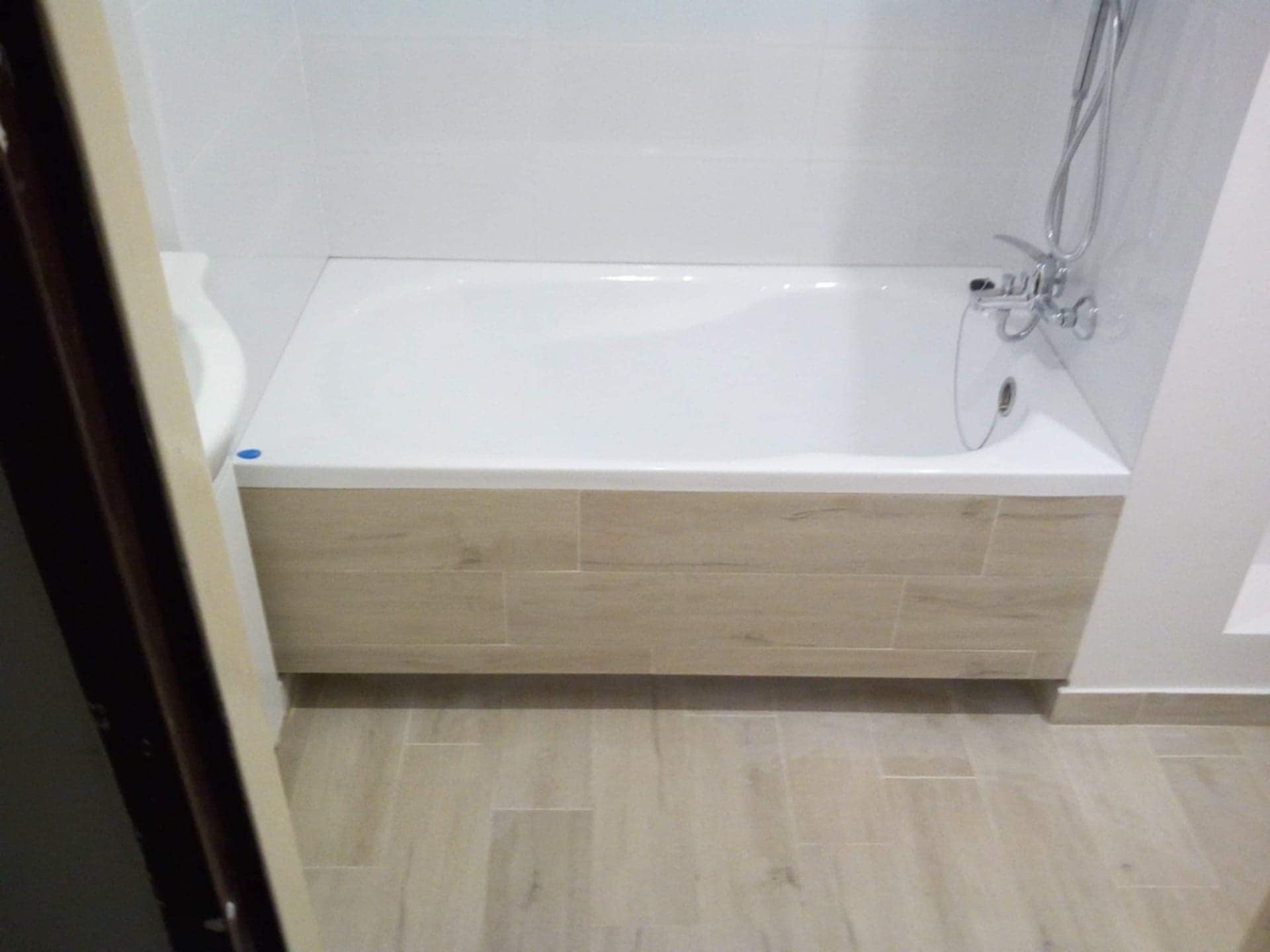 Biała wanna z obudową z płytek imitujących drewno w wykończonej łazience z jasnymi płytkami podłogowymi i ściennymi. Widoczna armatura.