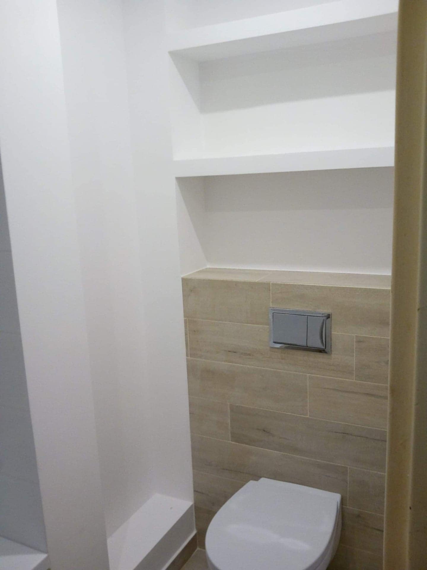 Wykończona toaleta w łazience z beżowymi płytkami imitującymi drewno, zabudowaną spłuczką i półkami wnękowymi. Minimalistyczny design, świeżo po remoncie.