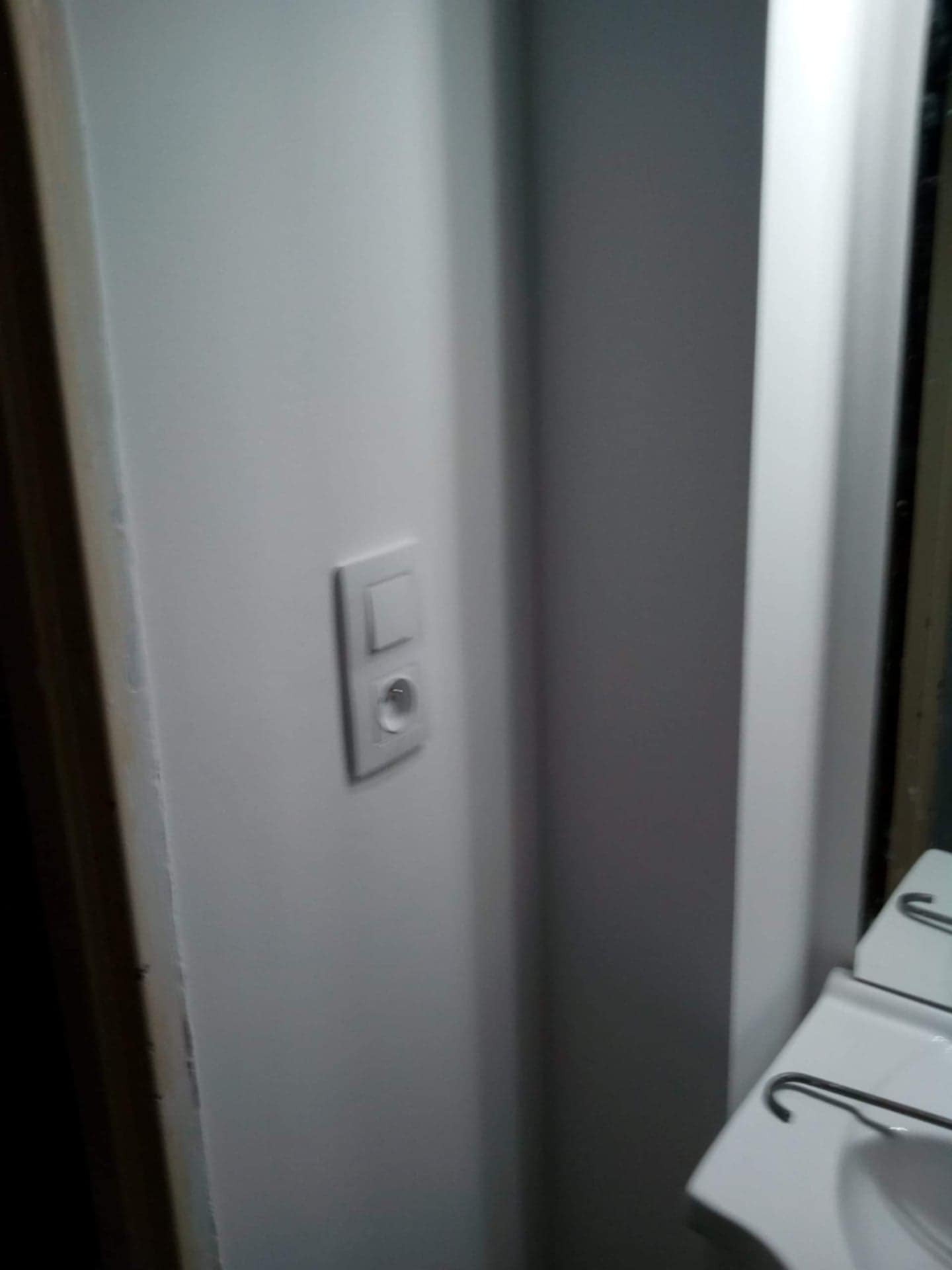 Biała ściana po malowaniu z gniazdkiem i włącznikiem, widoczny fragment umywalki i lustra. Minimalistyczne wykończenie łazienki.