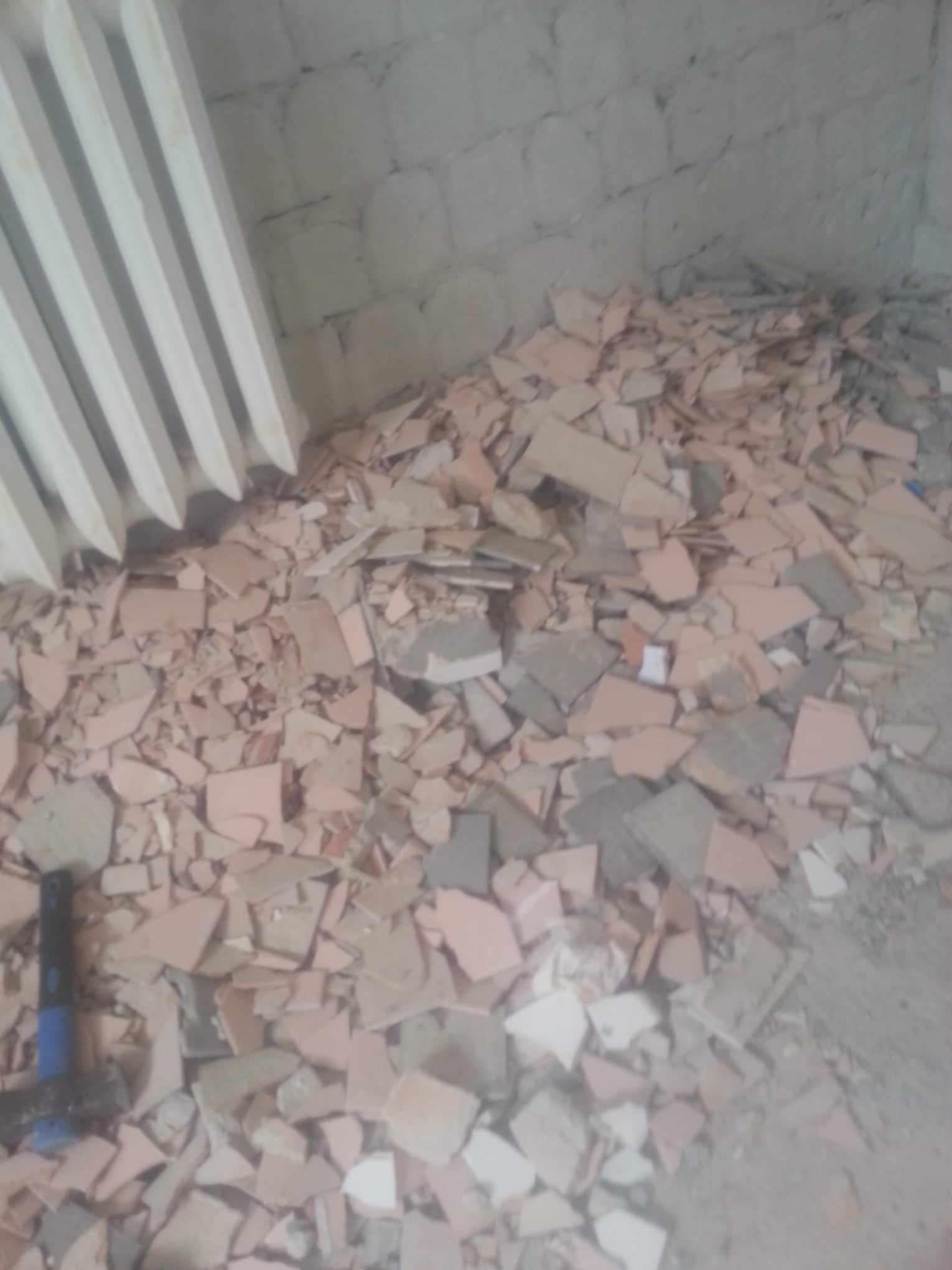 Stos potłuczonych, różowych i szarych płytek na podłodze w rogu pokoju, obok grzejnika i młotka. Widoczne ślady po demontażu płytek na ścianie.