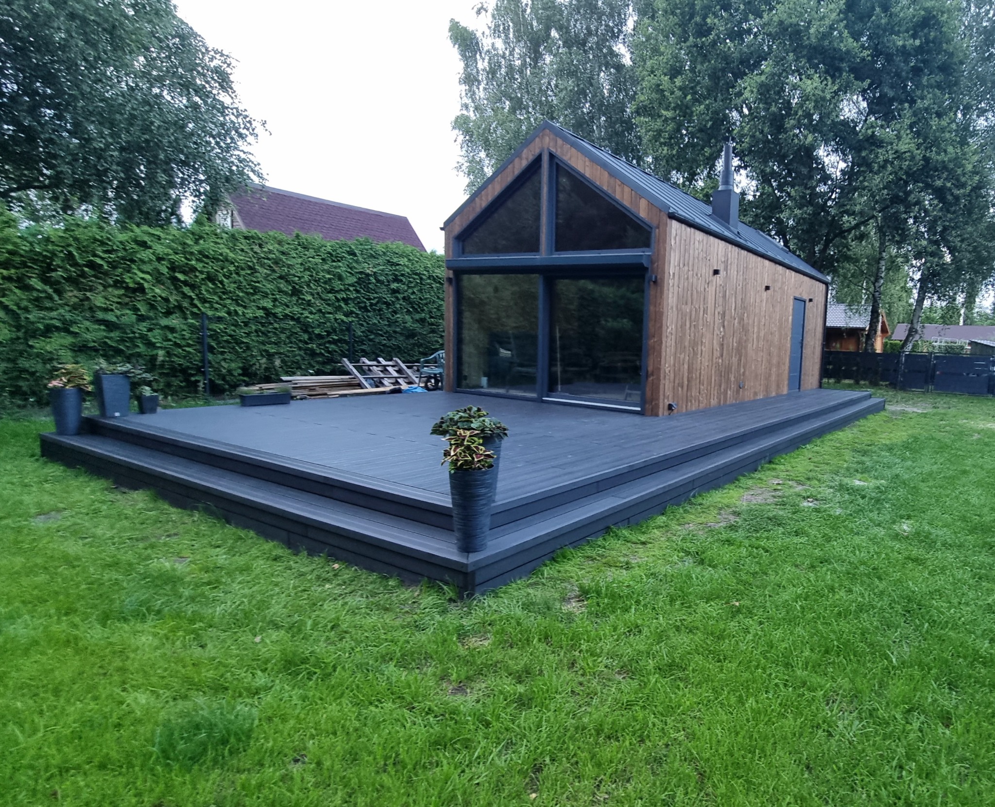 Nowoczesny, ciemnoszary taras kompozytowy z niskimi schodami, przylegający do drewnianego domku z dużymi oknami, otoczony zielonym trawnikiem i żywopłotem.
