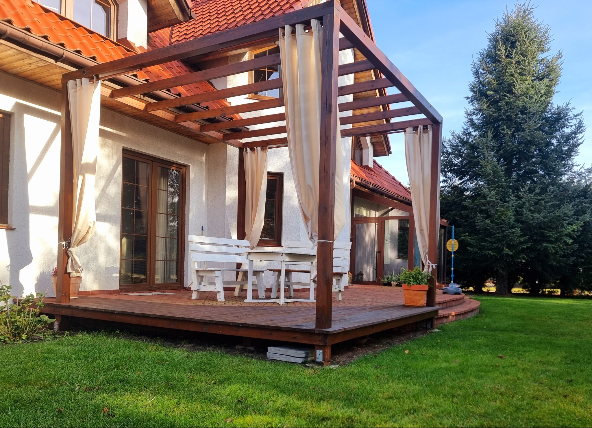 Drewniana konstrukcja z zasłonami na tarasie przy domu z czerwonym dachem. Na tarasie białe meble ogrodowe i donice z kwiatami. W tle zielony trawnik i drzewo.