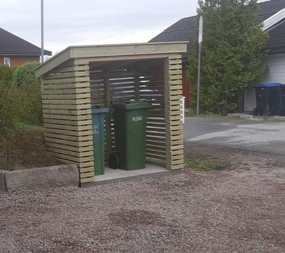 Drewniana konstrukcja osłaniająca dwa pojemniki na śmieci, z poziomymi listwami i skośnym dachem, na żwirowym podłożu, w tle domy jednorodzinne.