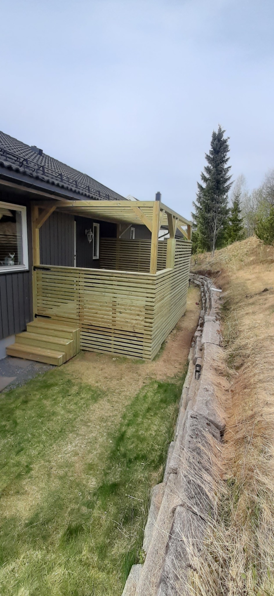 Nowoczesny taras z jasnego drewna, z ażurową balustradą i zadaszeniem, przylegający do domu z szarą elewacją. Widok z boku, na tle zieleni i drzew.