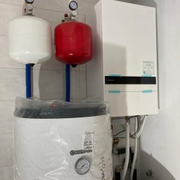 Ekociepło - Biały i czerwony zbiornik ciśnieniowy z manometrami, biały piec Panasonic oraz biały bojler Kospel SWP-200 w folii, zamontowane w pomieszczeniu z białymi płytkami.