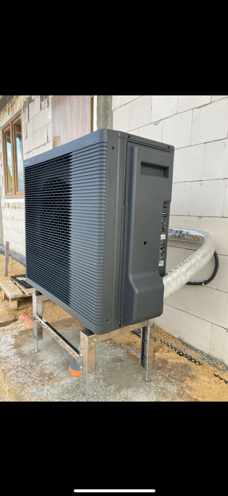 Szara pompa ciepła Aquarea na metalowym stelażu, zamontowana przy ścianie z białego bloczka. Widoczny fragment okna i rura w srebrnej izolacji.