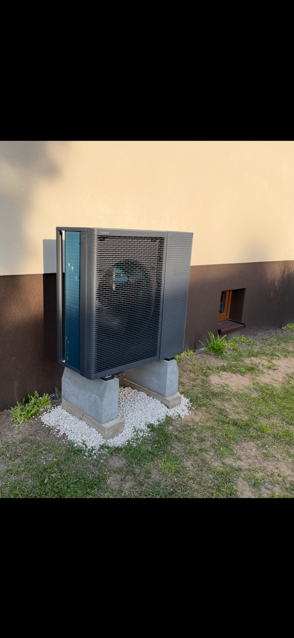 Zamontowana, szara pompa ciepła Panasonic na betonowych podstawach z białym żwirem, przy ścianie budynku z małym okienkiem piwnicznym i zieloną trawą.