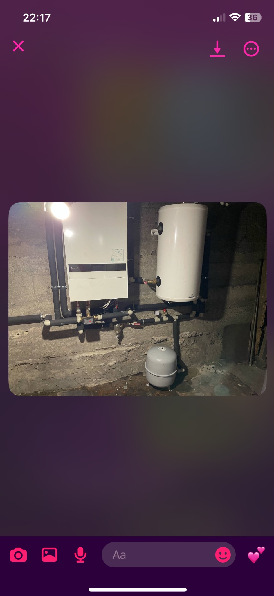 Biały piec CO wiszący na szarej, betonowej ścianie piwnicy, z podłączonymi czarnymi rurami i białym zbiornikiem na wodę. Widoczny szary zbiornik wyrównawczy.