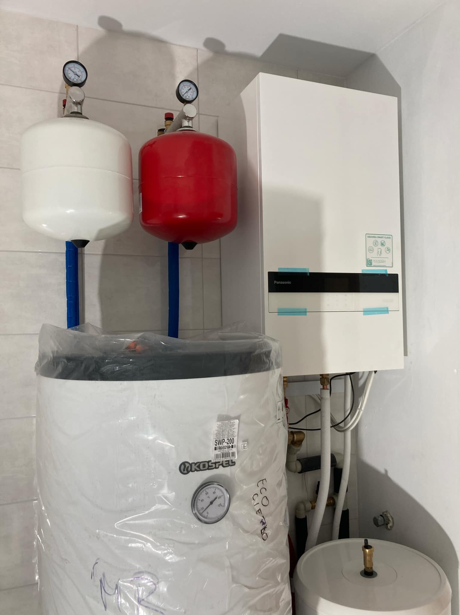 Biały i czerwony zbiornik ciśnieniowy z manometrami, biały piec Panasonic oraz biały bojler Kospel SWP-200 w folii, zamontowane w pomieszczeniu z białymi płytkami.