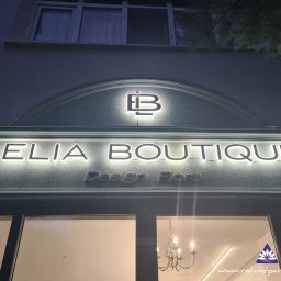 Region Projekt Weronika Galewska - Podświetlany szyld butiku 'Lelia Boutique Design Floral' z logo nad wejściem, widoczny z zewnątrz, oświetlony wieczorową porą. W tle okno i fragment wnętrza.