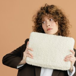 Region Projekt Weronika Galewska - Kobieta z burzą loków w brązowej marynarce trzymająca kremową, futerkową torbę na beżowym tle. Spojrzenie skierowane w lewo, delikatny makijaż.