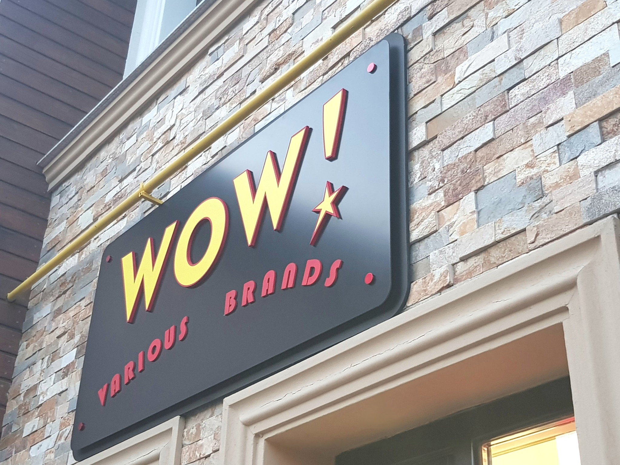 Elegancki szyld reklamowy 'WOW! Various Brands' z żółto-czerwonym napisem, zamontowany na kamiennej elewacji budynku, widoczny z dołu pod kątem.