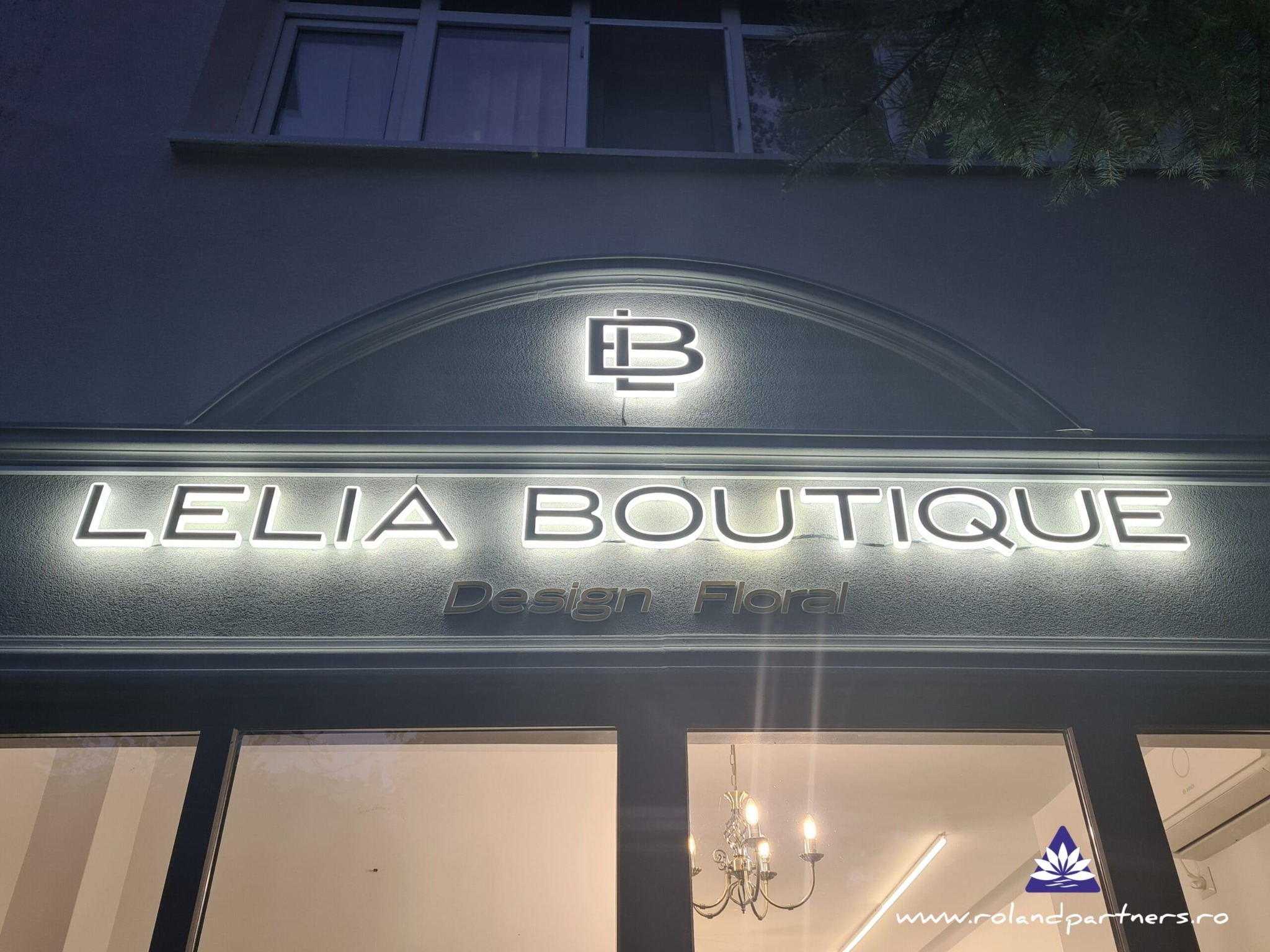Podświetlany szyld butiku 'Lelia Boutique Design Floral' z logo nad wejściem, widoczny z zewnątrz, oświetlony wieczorową porą. W tle okno i fragment wnętrza.