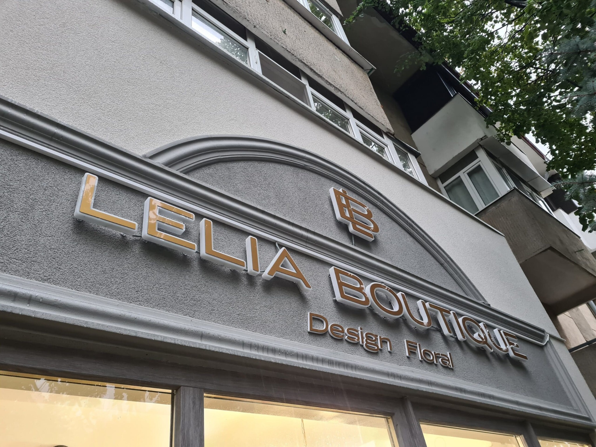 Elegancki szyld sklepu 'Lelia Boutique Design Floral' z podświetlanymi literami w kolorze złota, umieszczony na szarej fasadzie budynku z dekoracyjnymi elementami.