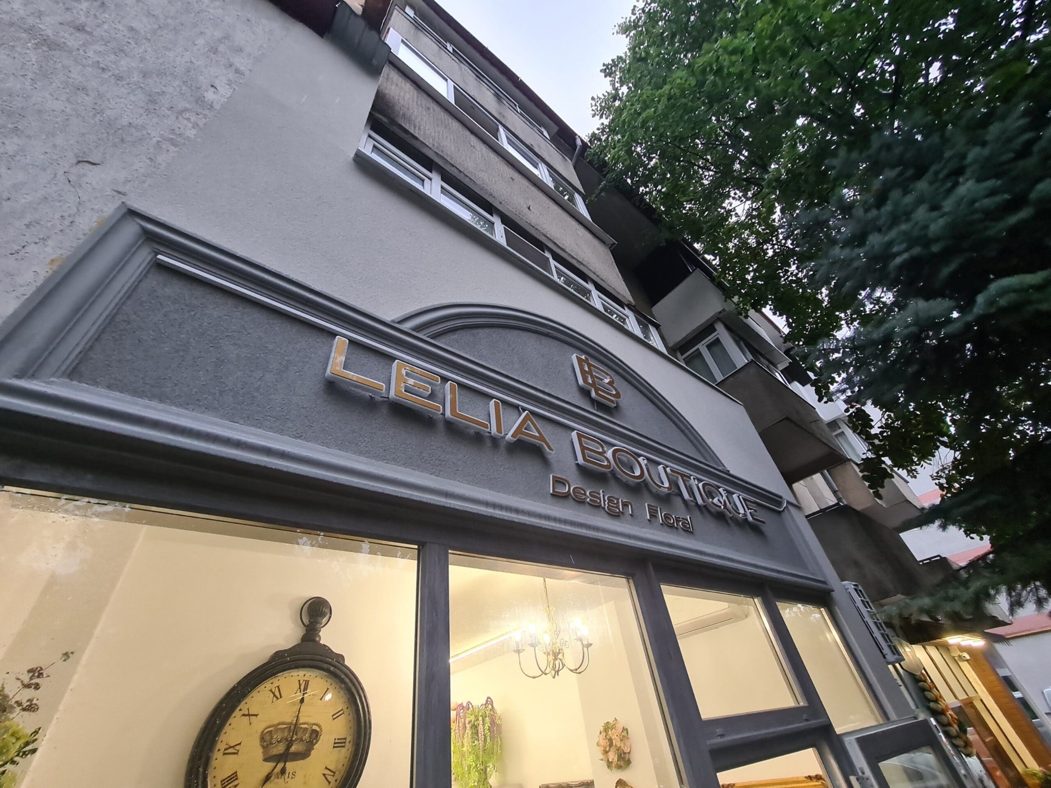 Fasada budynku z szyldem 'Lelia Boutique Design Floral', widok z dołu. W oknie widoczny zegar i żyrandol, a w tle fragmenty zieleni drzew.