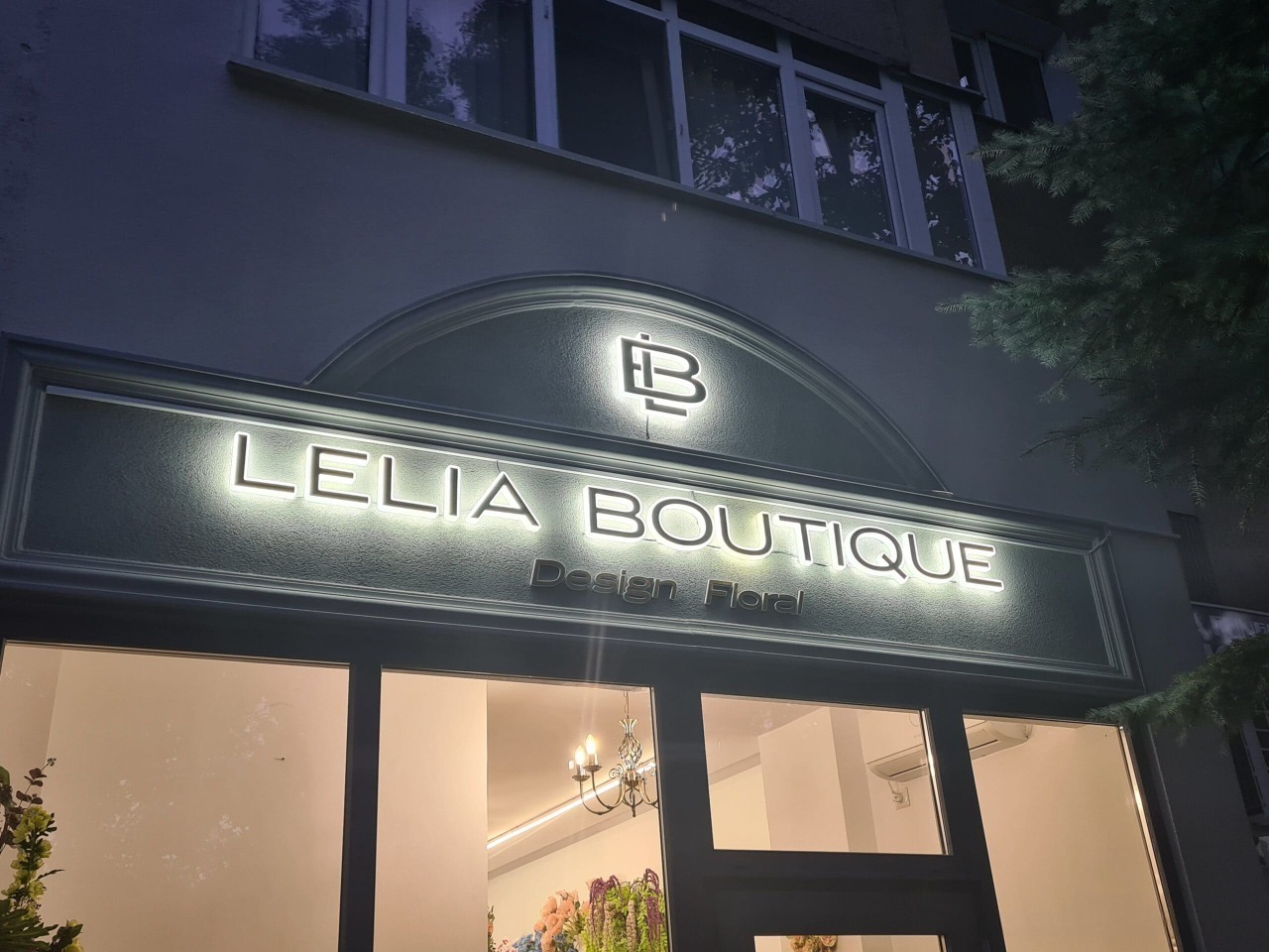 Podświetlany szyld sklepu 'Lelia Boutique Design Floral' z logo nad wejściem, widoczny z zewnątrz, z elementami roślinnymi w oknach i oświetleniem wewnętrznym.
