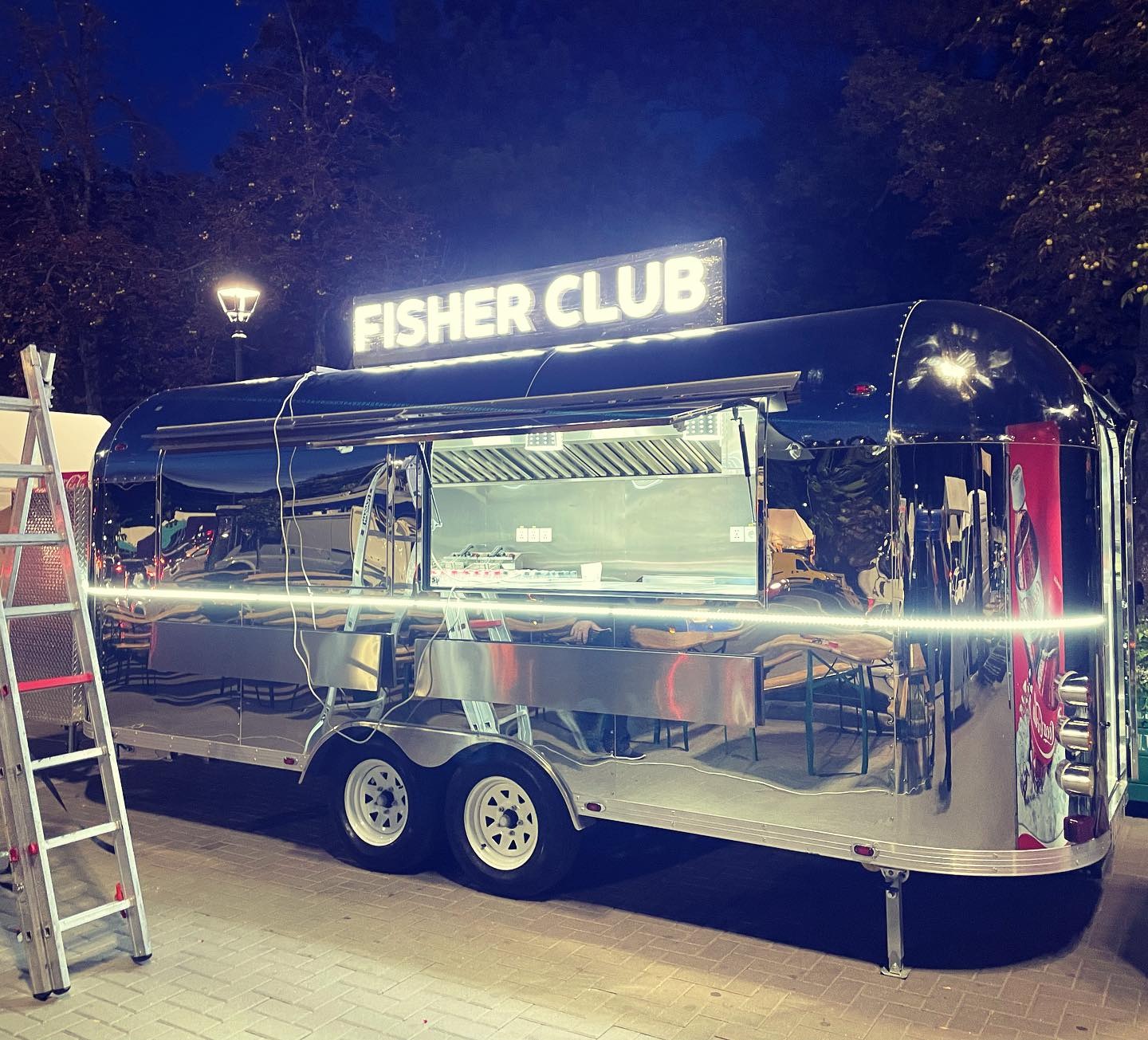 Błyszcząca, metalowa przyczepa gastronomiczna 'Fisher Club' z oświetlonym szyldem, zaparkowana na zewnątrz, widoczna drabina z boku. Nocne ujęcie.