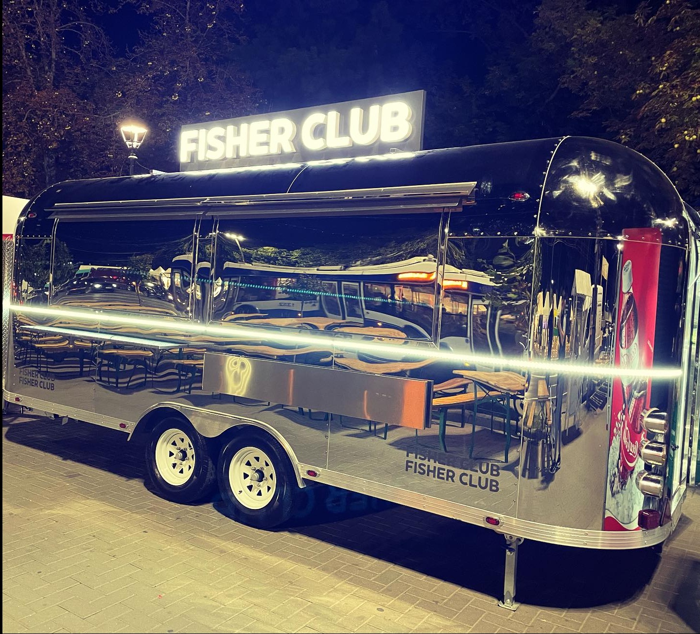 Lśniąca, metalowa przyczepa gastronomiczna 'Fisher Club' z podświetlanym szyldem, odbijająca otoczenie w polerowanej powierzchni, ustawiona na zewnątrz w nocy.