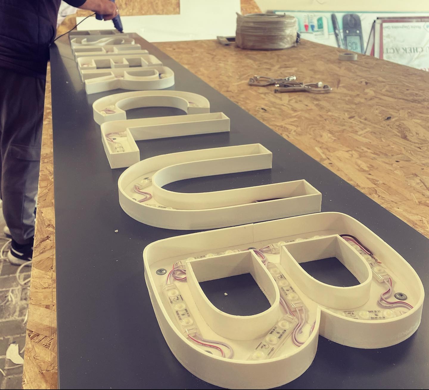 Montaż podświetlanych liter przestrzennych na płycie. Widoczne diody LED wewnątrz liter i narzędzia pracy. Etap produkcji szyldu reklamowego.