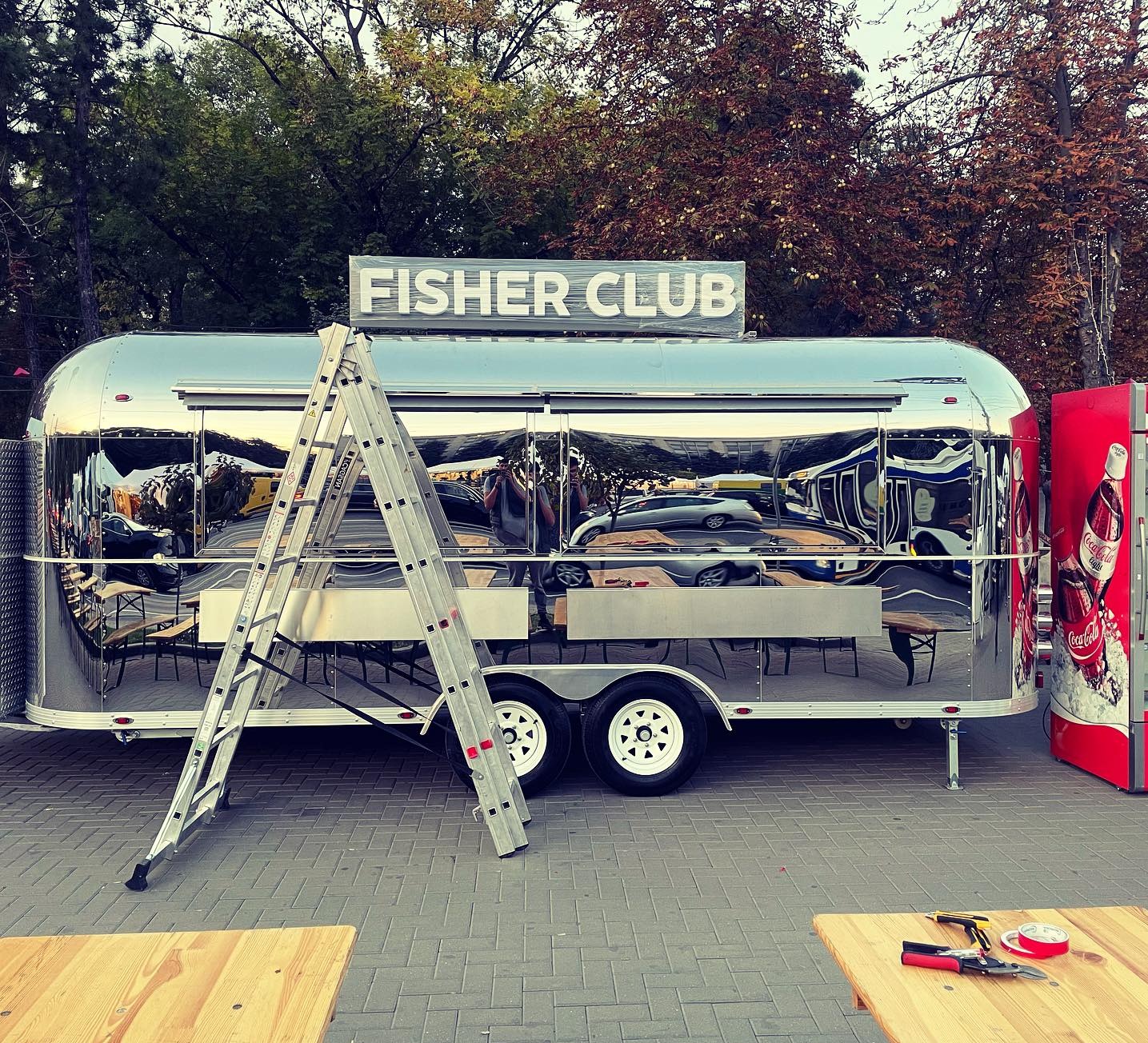 Błyszcząca, metalowa przyczepa gastronomiczna 'Fisher Club' z drabiną opartą o bok, sugerująca montaż lub prace wykończeniowe. W tle drzewa i automat Coca-Cola.