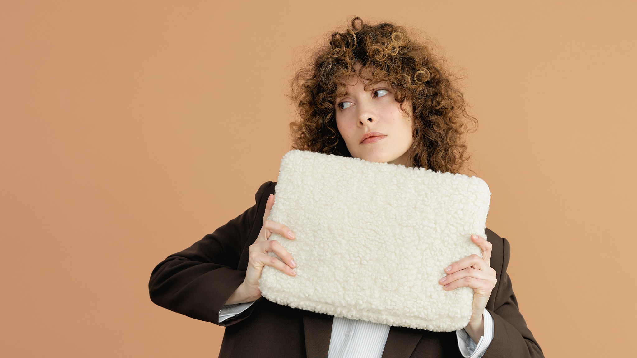 Kobieta z burzą loków w brązowej marynarce trzymająca kremową, futerkową torbę na beżowym tle. Spojrzenie skierowane w lewo, delikatny makijaż.