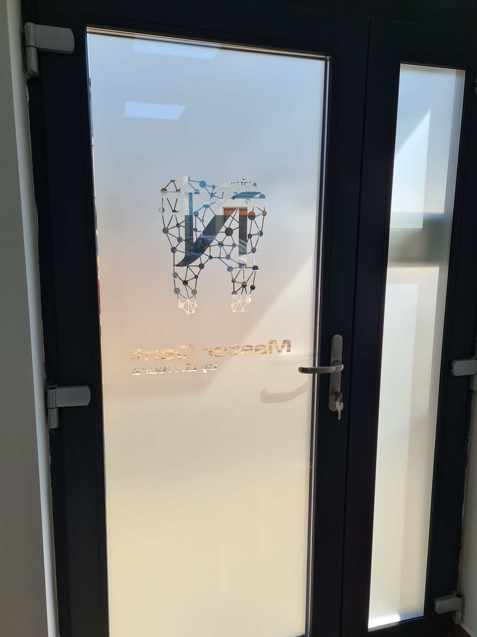 Szklane drzwi z naklejonym logo dentystycznym w formie zęba połączonego siecią, z napisem poniżej. Matowa folia na szkle zapewnia prywatność.
