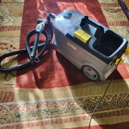 Wypierz to - Maszyna Karcher do prania dywanów stoi na wzorzystym dywanie w odcieniach czerwieni i beżu, gotowa do użycia. Widoczne detale urządzenia i faktura dywanu.