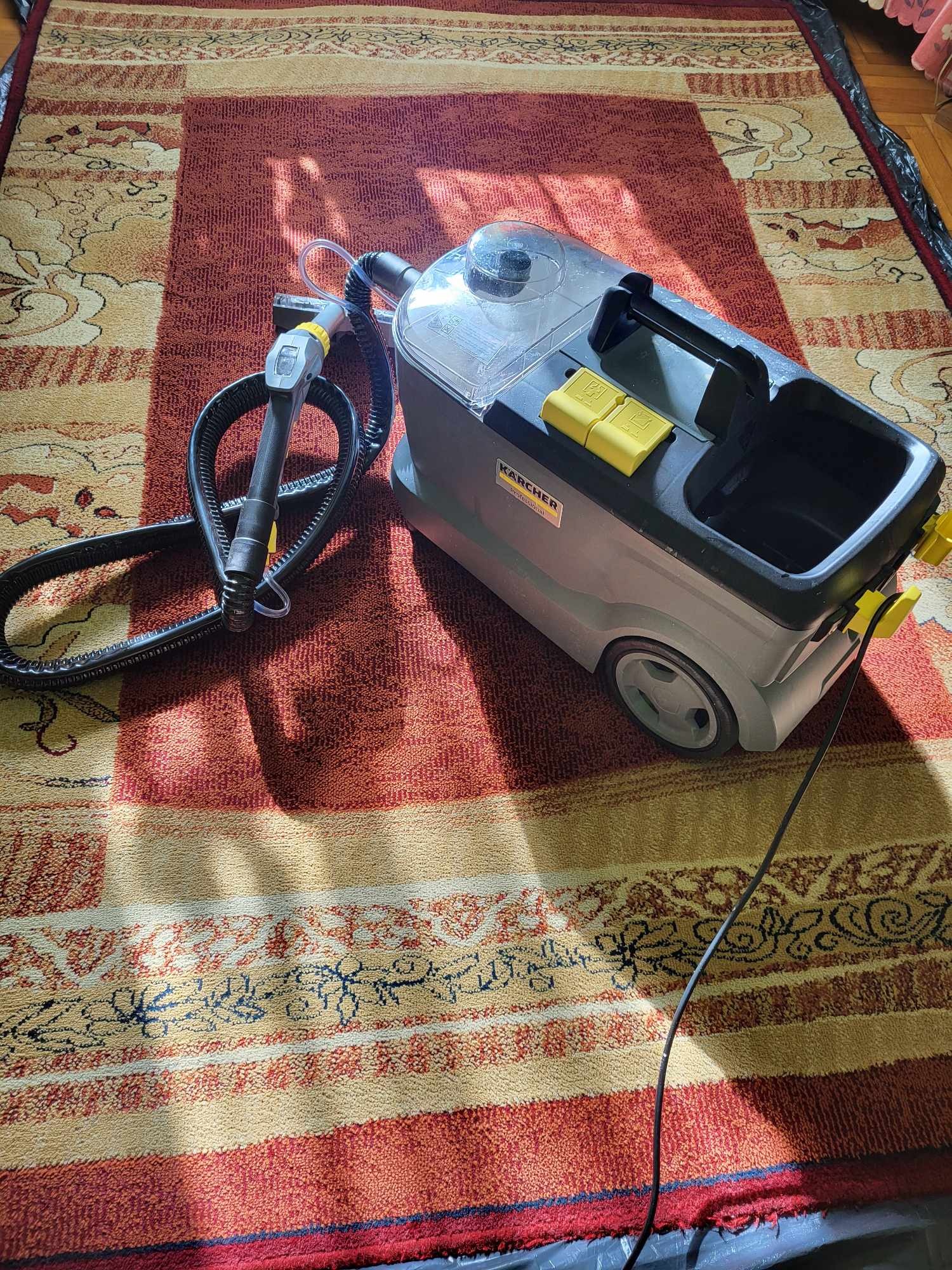 Maszyna Karcher do prania dywanów stoi na wzorzystym dywanie w odcieniach czerwieni i beżu, gotowa do użycia. Widoczne detale urządzenia i faktura dywanu.