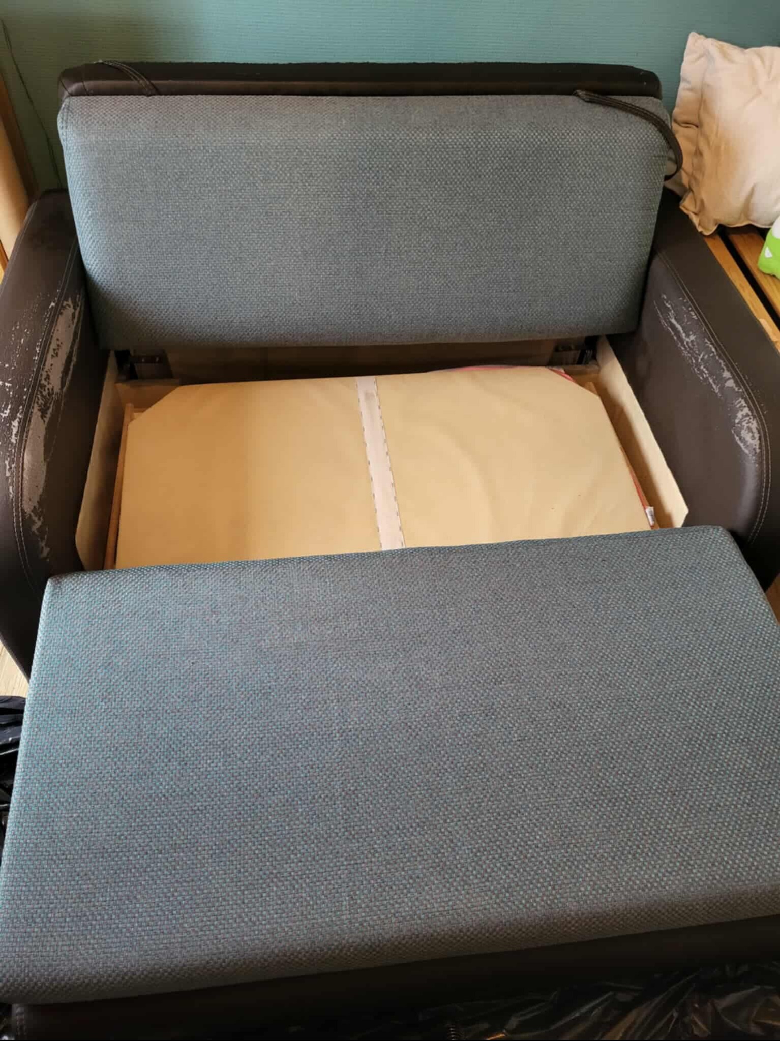 Rozłożona kanapa z widocznym pojemnikiem na pościel i zużytą tapicerką na bokach. Widoczne ślady użytkowania i zabrudzenia na materiale.