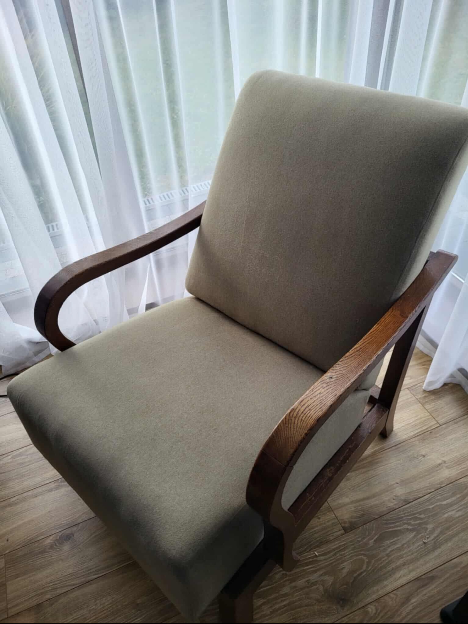 Fotel tapicerowany w odcieniach beżu z drewnianymi podłokietnikami, po czyszczeniu, na tle okna z białymi zasłonami. Widok z góry, naturalne światło.