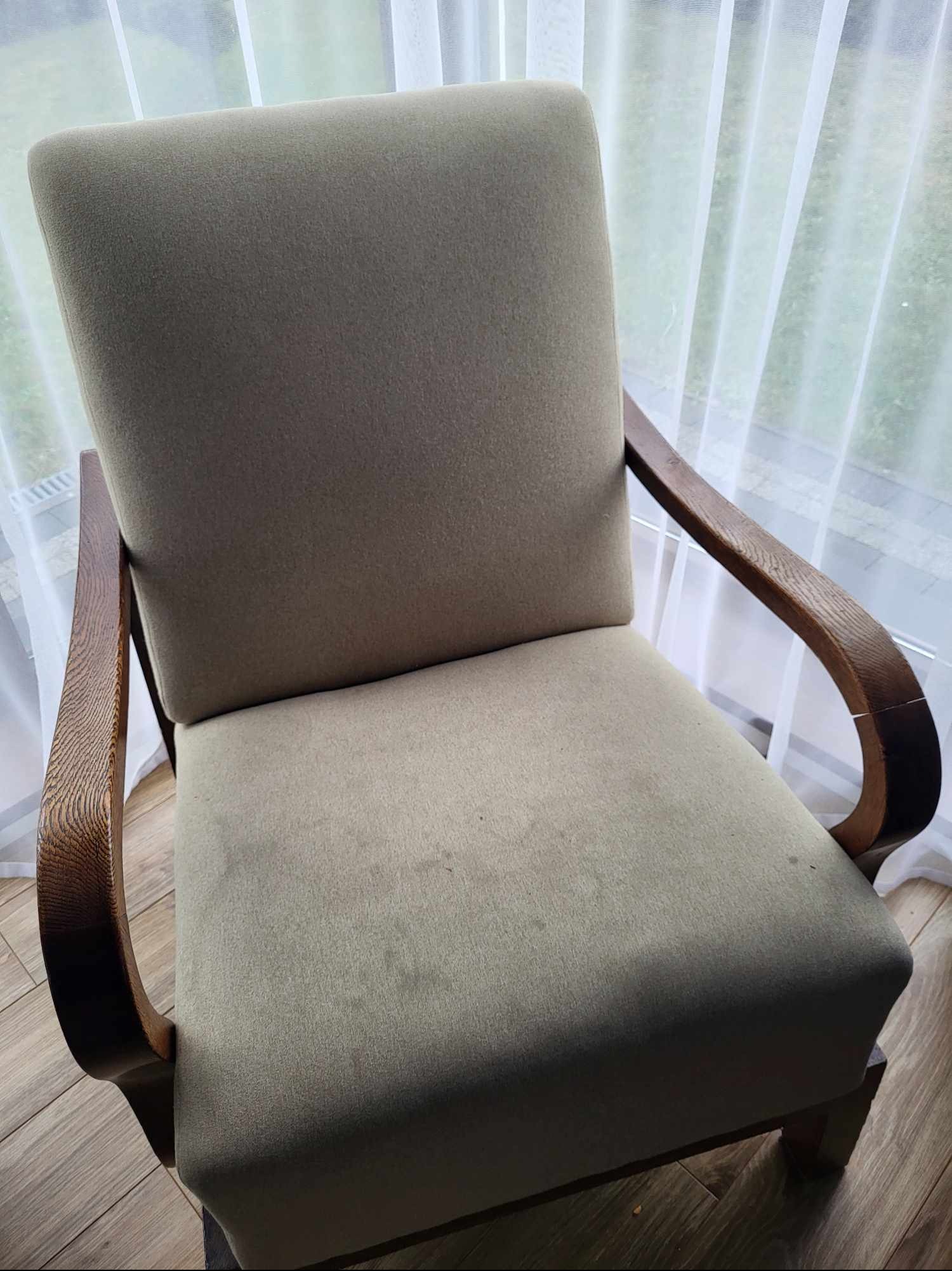 Fotel z beżową tapicerką i drewnianymi podłokietnikami, widoczne plamy na siedzisku, na tle okna z firanką. Fotografia 'przed' czyszczeniem.