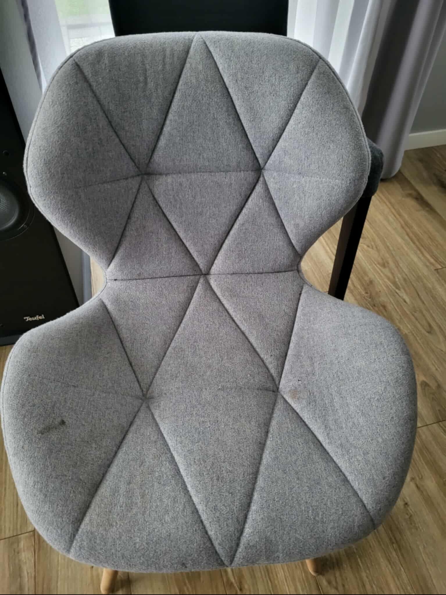 Szare krzesło tapicerowane z geometrycznym wzorem przed praniem. Widoczne plamy na siedzisku, drewniana podłoga i fragment głośnika w tle.