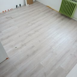 Sowbud Cezary Sowiński - Jasne panele podłogowe ułożone w pokoju, widoczne listwy przypodłogowe i grzejnik w kolorze zielonym. Fragment ściany z zamontowanymi elementami instalacji.