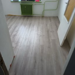 Sowbud Cezary Sowiński - Jasne panele podłogowe ułożone w pokoju z zielonym grzejnikiem i rurami. Widoczne fragmenty drzwi i okna. Ujęcie z poziomu podłogi.