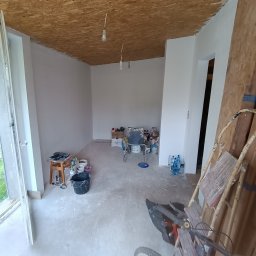Sowbud Cezary Sowiński - Wnętrze pokoju w trakcie malowania ścian na biało. Widoczne narzędzia malarskie, drabina i surowy strop z płyty OSB. Częściowo otwarte drzwi balkonowe.