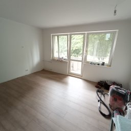 Sowbud Cezary Sowiński - Jasne panele podłogowe w pokoju z białymi ścianami i oknem balkonowym. Widoczny odkurzacz przemysłowy i narzędzia, sugerujące prace wykończeniowe.