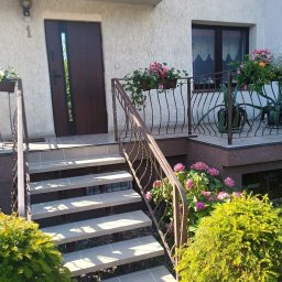 Sowbud Cezary Sowiński - Wejście do domu z brązowymi drzwiami i schodami wyłożonymi płytkami. Balustrada z metalu, donice z kwiatami i krzewy ozdobne.