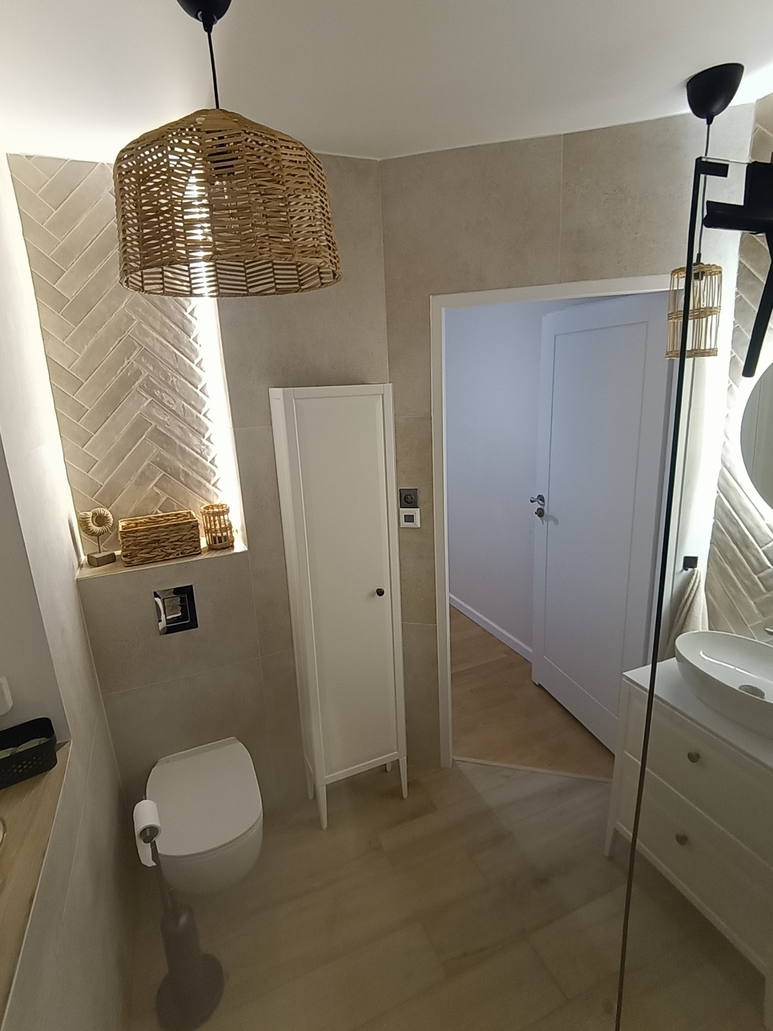 Jasna łazienka z beżowymi płytkami ułożonymi w jodełkę, wiklinową lampą i białymi meblami. Widoczne WC, szafka, umywalka i drzwi do innego pomieszczenia.