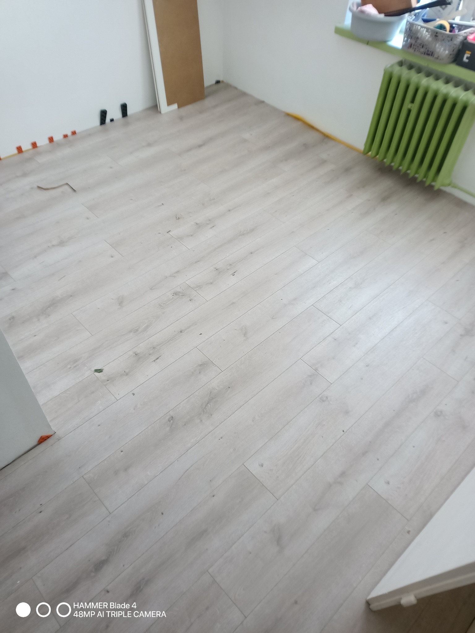 Jasne panele podłogowe ułożone w pokoju, widoczne listwy przypodłogowe i grzejnik w kolorze zielonym. Fragment ściany z zamontowanymi elementami instalacji.