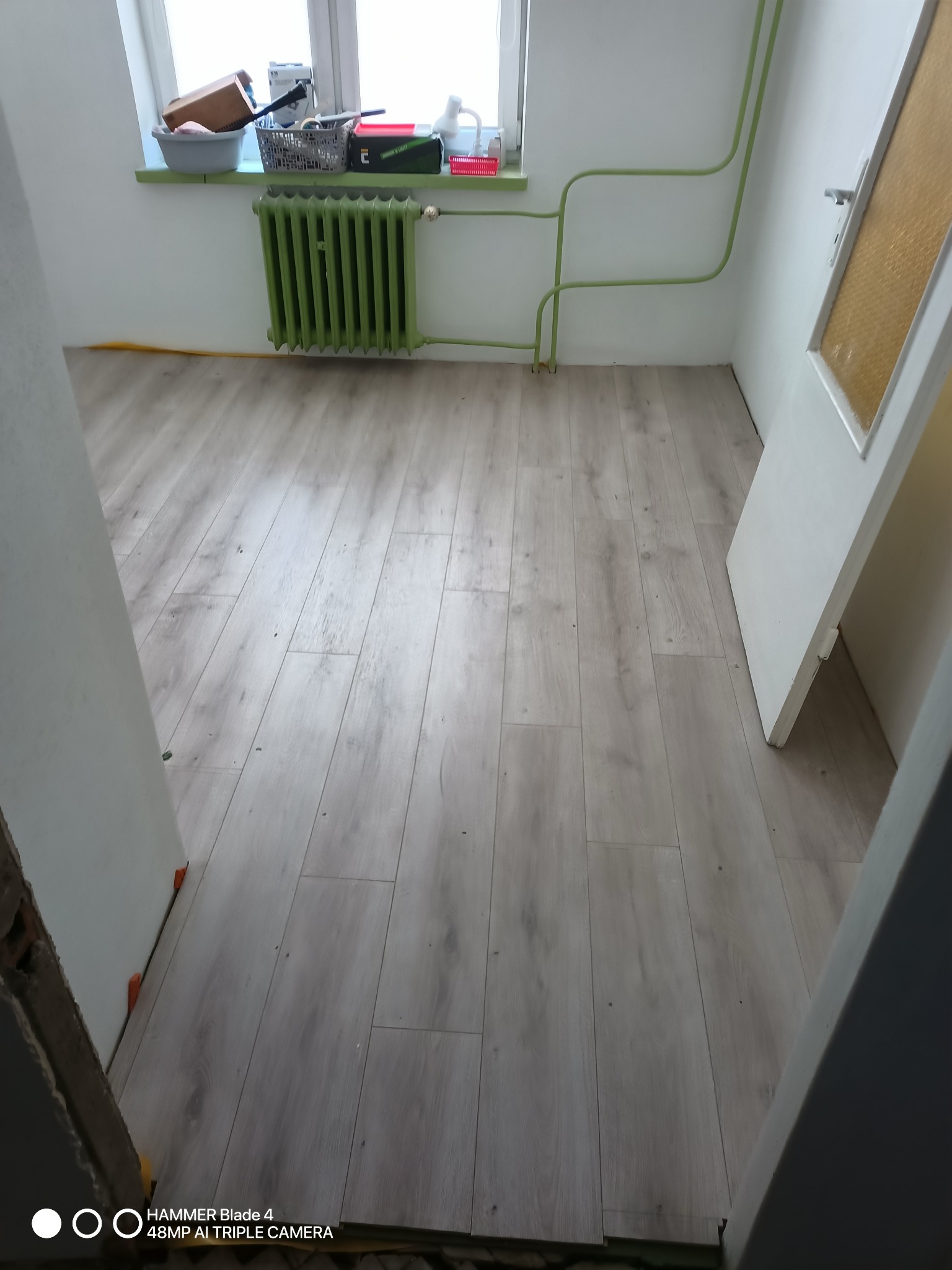 Jasne panele podłogowe ułożone w pokoju z zielonym grzejnikiem i rurami. Widoczne fragmenty drzwi i okna. Ujęcie z poziomu podłogi.