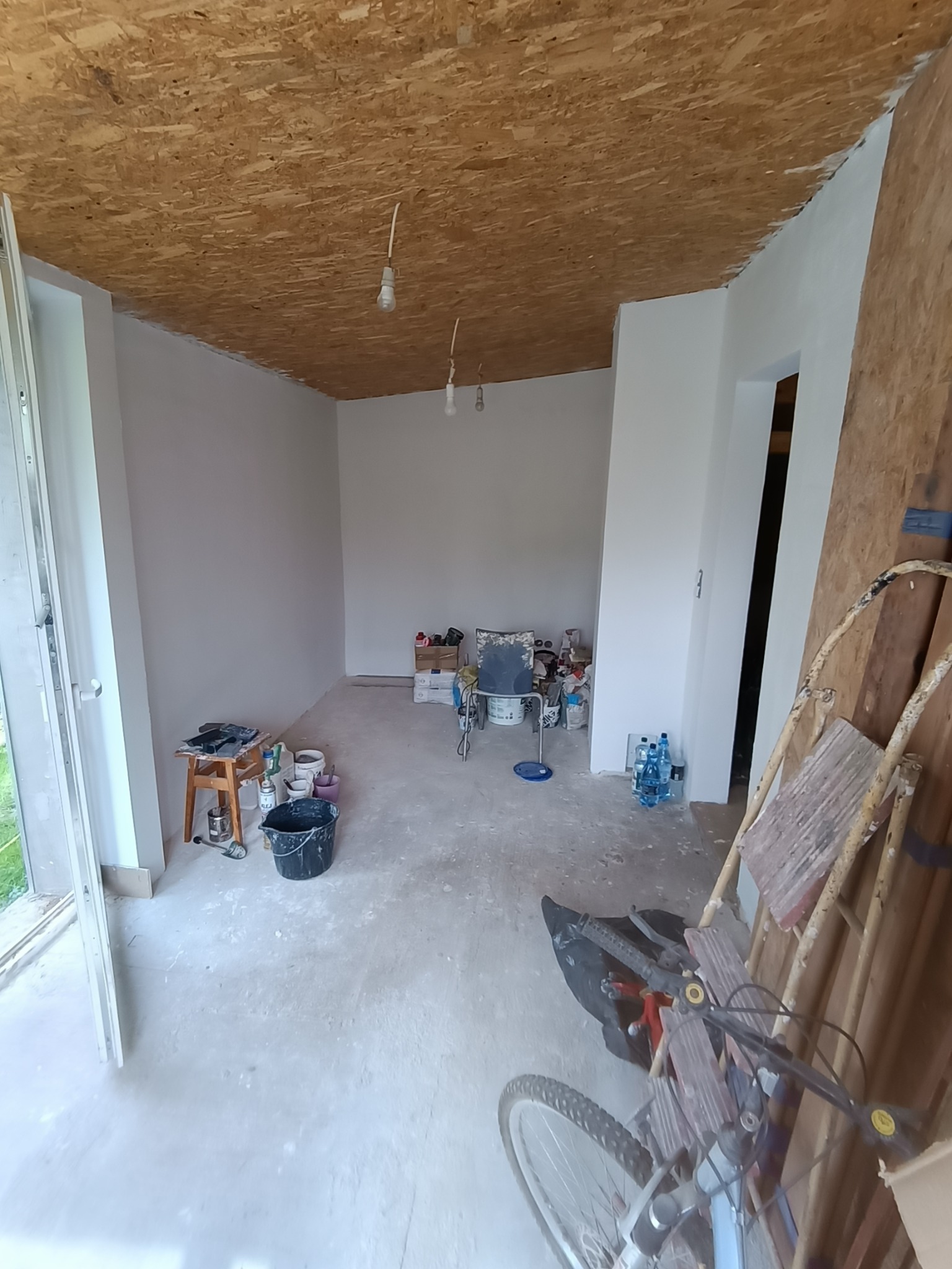 Wnętrze pokoju w trakcie malowania ścian na biało. Widoczne narzędzia malarskie, drabina i surowy strop z płyty OSB. Częściowo otwarte drzwi balkonowe.