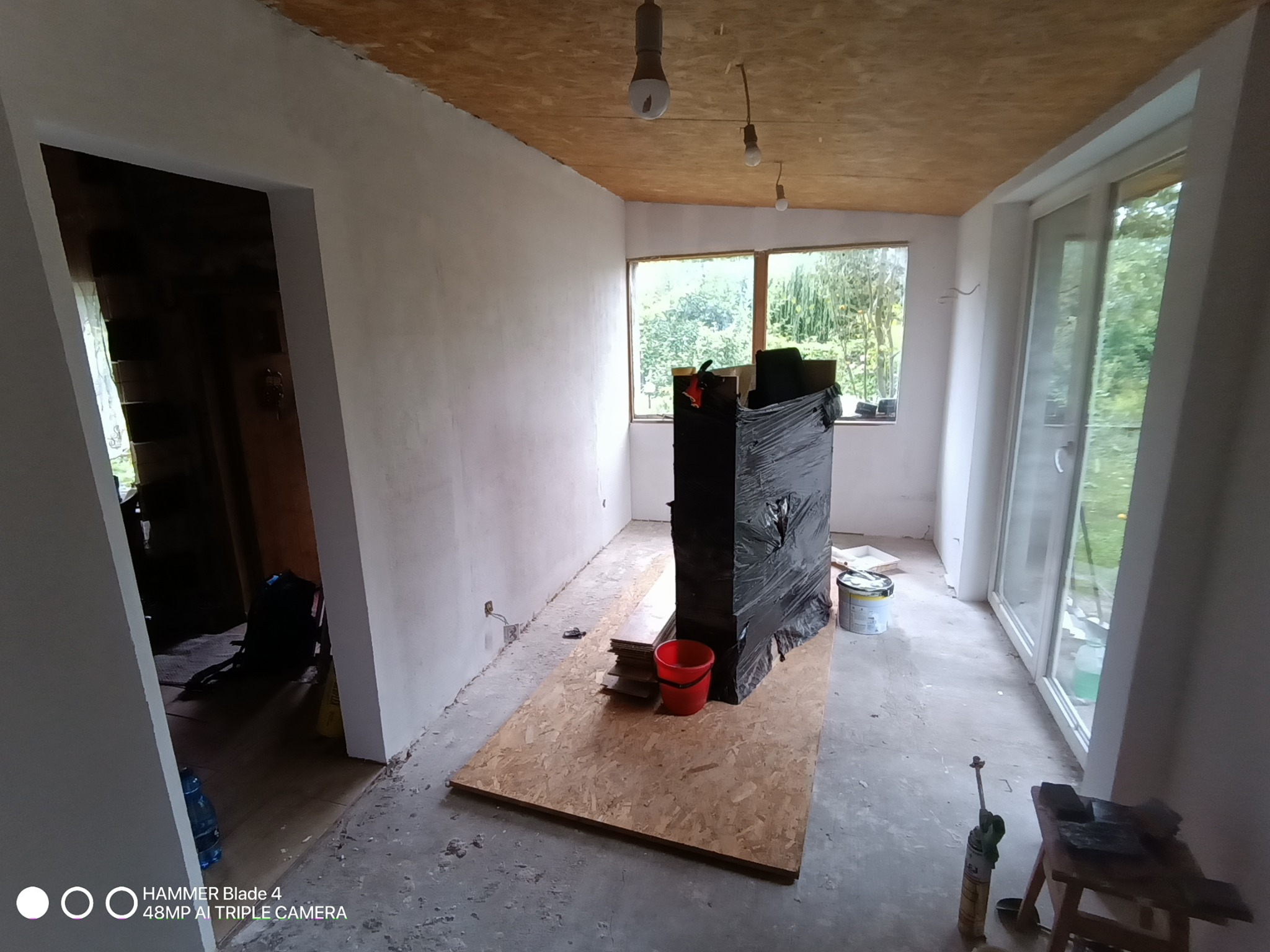 Wnętrze w trakcie prac wykończeniowych: surowe ściany, okno z widokiem na zieleń, zabezpieczony kominek, płyta OSB na podłodze, narzędzia i materiały budowlane.