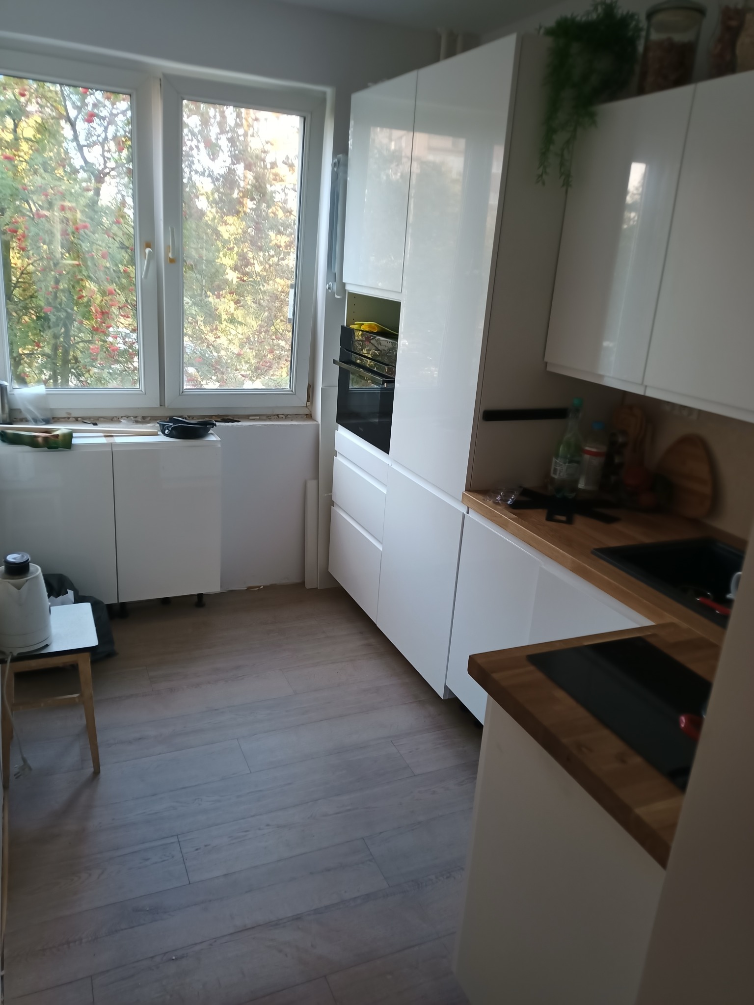 Jasna kuchnia w trakcie montażu: białe szafki, drewniany blat, okno z widokiem na drzewa. Widoczne niedokończone prace przy oknie i podłodze.