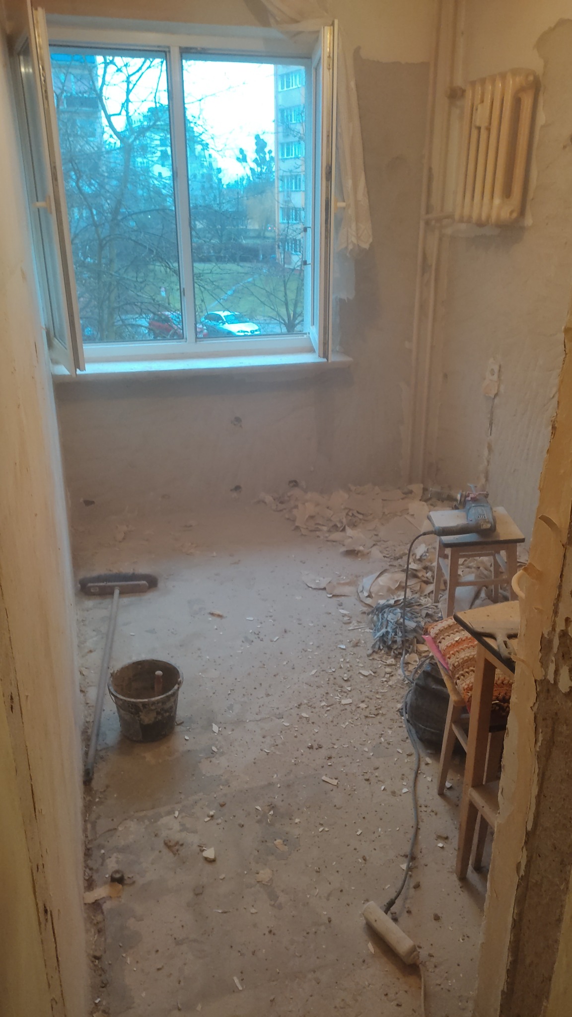 Pokój w trakcie remontu: gołe ściany, resztki tapet na podłodze, drabina, wiadro i narzędzia. Widok z okna na blok mieszkalny. Etap przygotowania do malowania.
