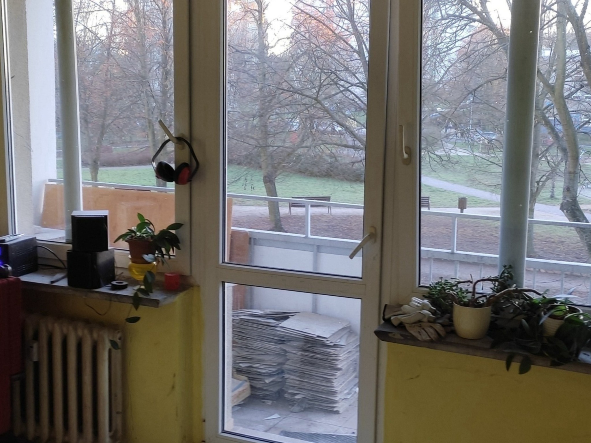 Balkon przed remontem: stos płytek, słuchawki na haku, rośliny doniczkowe na parapecie. Widok na park przez okno. Przygotowania do prac wykończeniowych.
