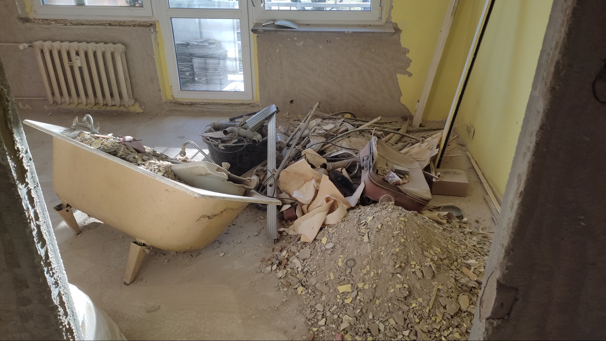 Wnętrze w trakcie demontażu: stara wanna wypełniona gruzem, widoczne fragmenty ścian bez płytek, rury i inne elementy instalacji. Przygotowanie do remontu łazienki.